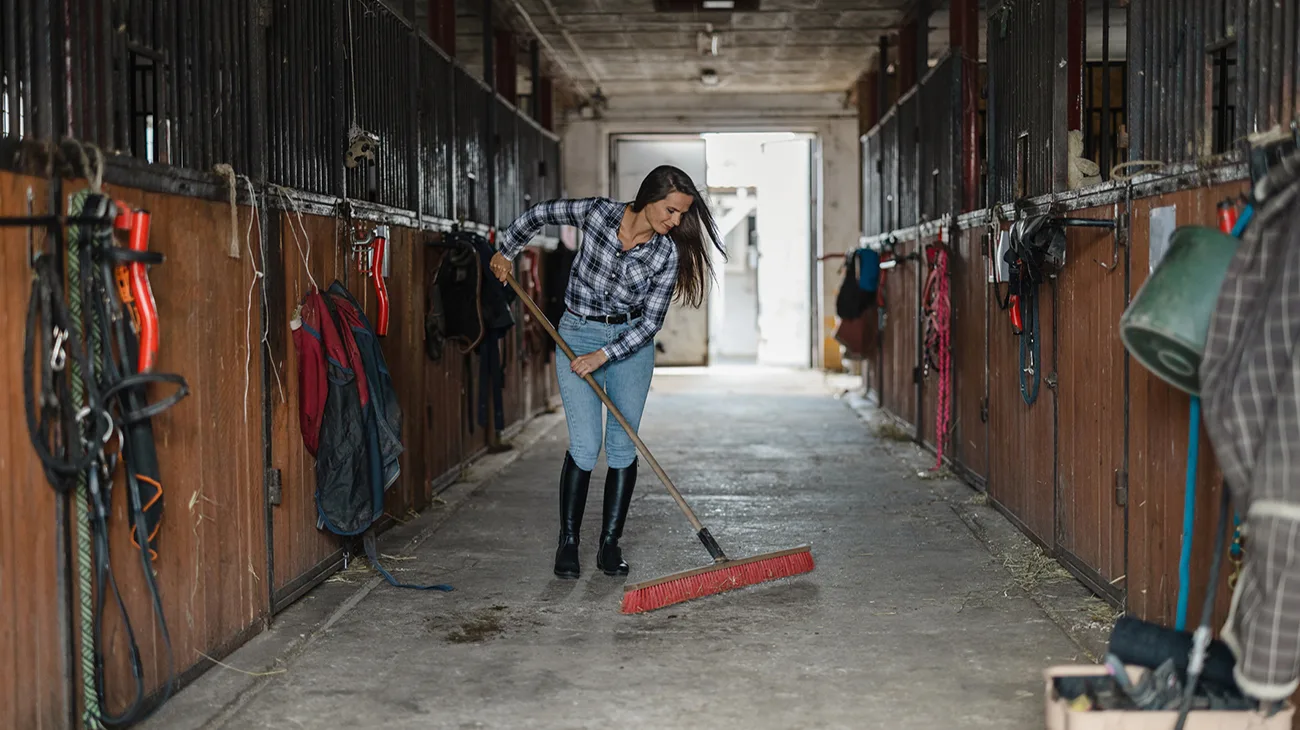 Young woman sweeping stable's floor with a broom